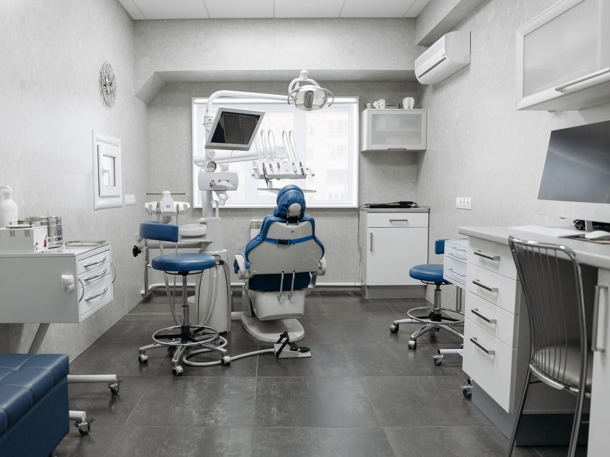 A treatment room with a blue dental chair, stools and storage cabinets